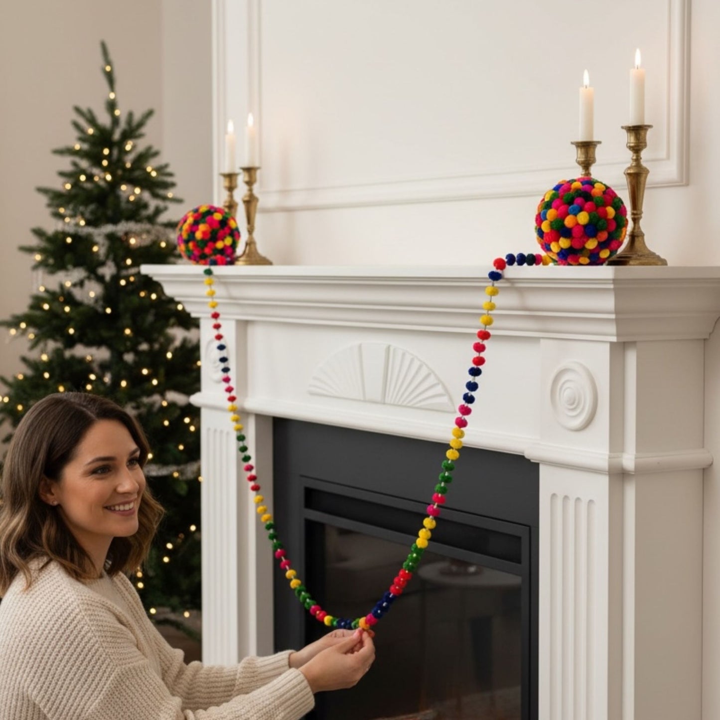 A woman hangs a colorful pom-pom garland draped over a white fireplace mantel in a decorated living room with a Christmas tree and lit candles.