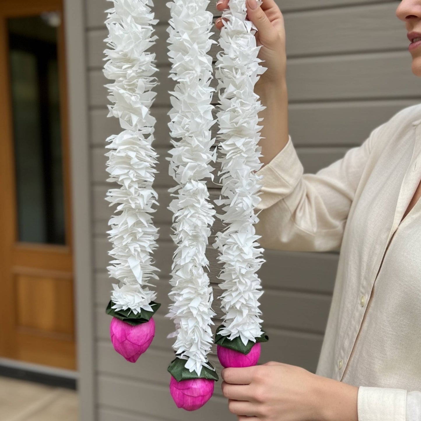 Close-up of two white artificial jasmine garland strands held up by a person outdoors, showing detailed petal texture with deep pink lotus bud finials and green leaf accents at the ends