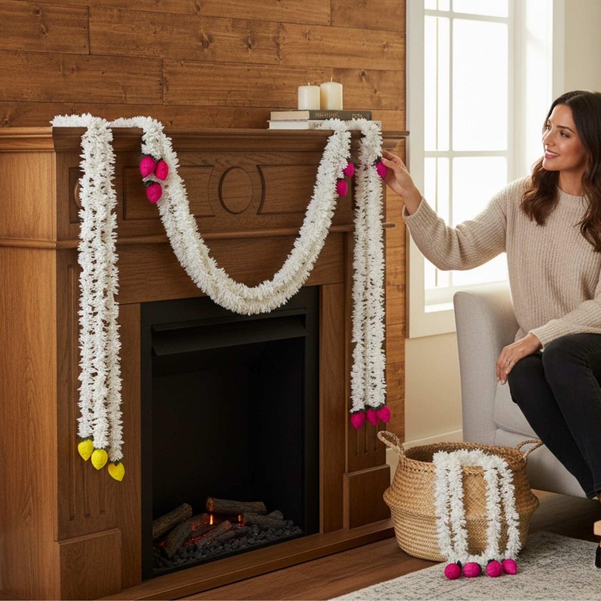 Woman arranging white jasmine artificial floral garlands draped over a wooden fireplace mantel, with pink and yellow lotus bud ends, and a wicker basket holding additional garlands on the floor