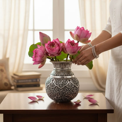 Person arranging pink lotus flowers in an ornate silver vase or kalash or lota on a table.