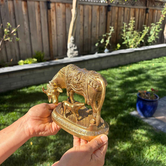 Hands holding Brass Kamdhenu Cow and Calf Idol outdoors, showcasing antique gold finish