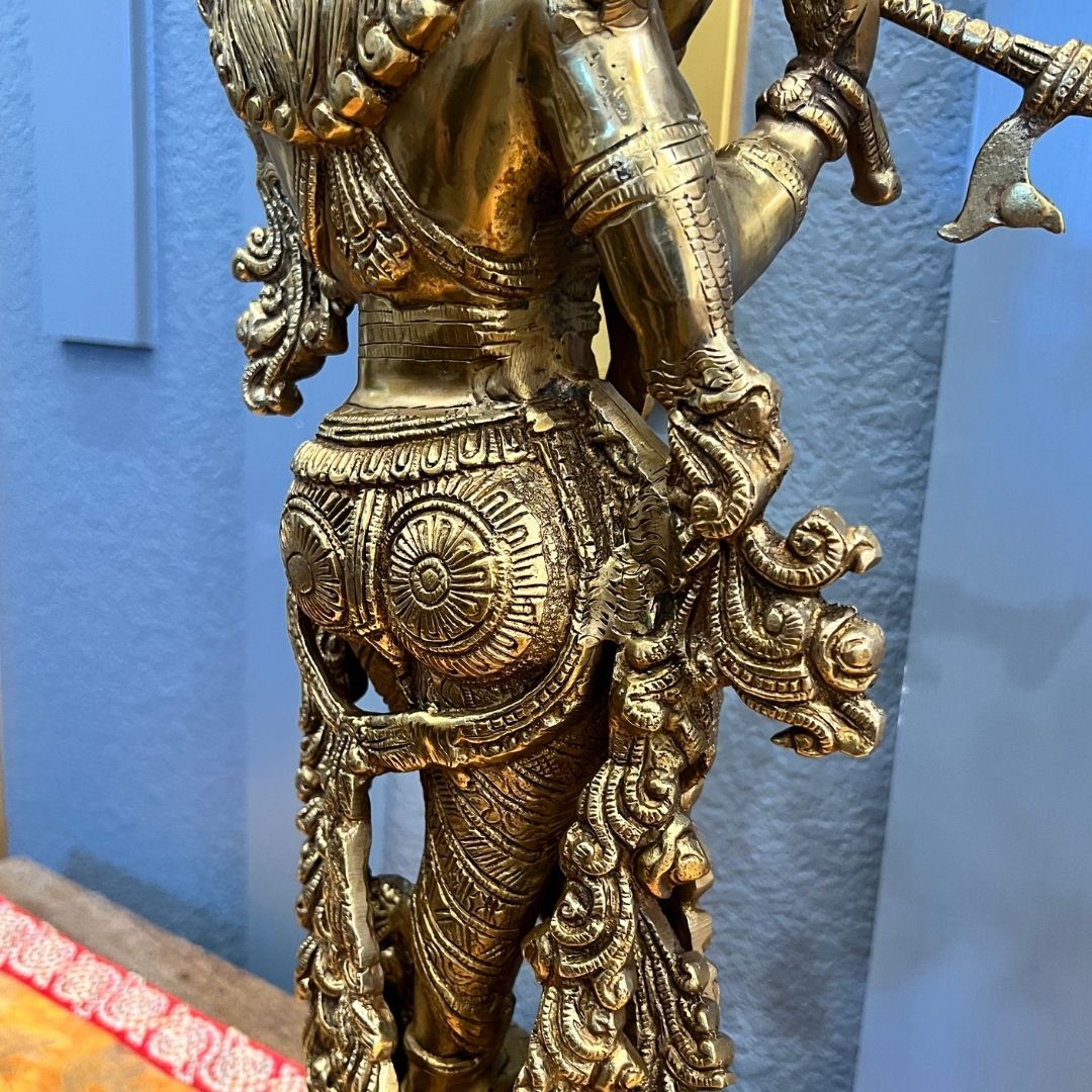 Back and side view of the brass Krishna statue showing hand-engraved detailing across the dhoti, sash and halo disc, against a blue wall