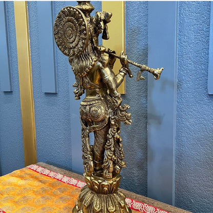 Three-quarter back view of the brass Krishna statue showing the flute, engraved jewellery and the circular lotus-mandala halo disc behind the head, against a blue paneled wall