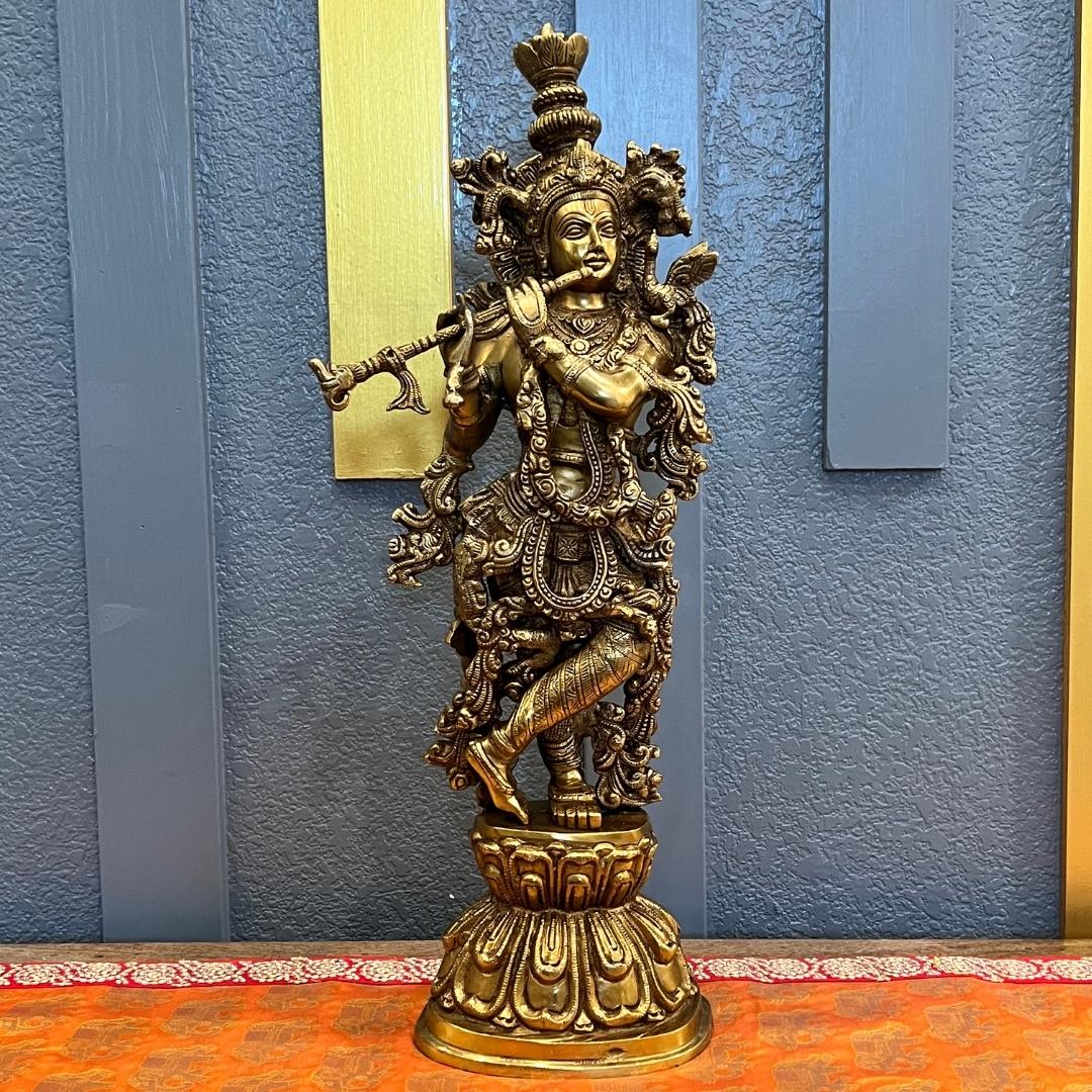 Brass Krishna statue standing in tribhanga posture on a layered lotus base, against a blue textured wall with gold vertical panels