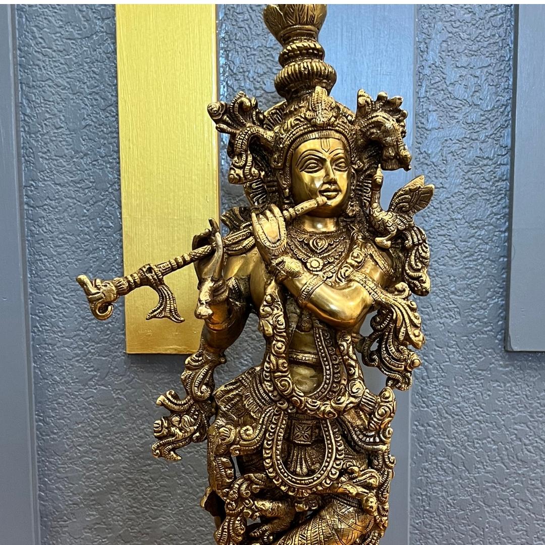 Close-up of the 24 inch brass Krishna statue, showing the tiered crown, engraved headdress and the bansuri flute held to his lips, against a gray textured wall with a gold vertical panel
