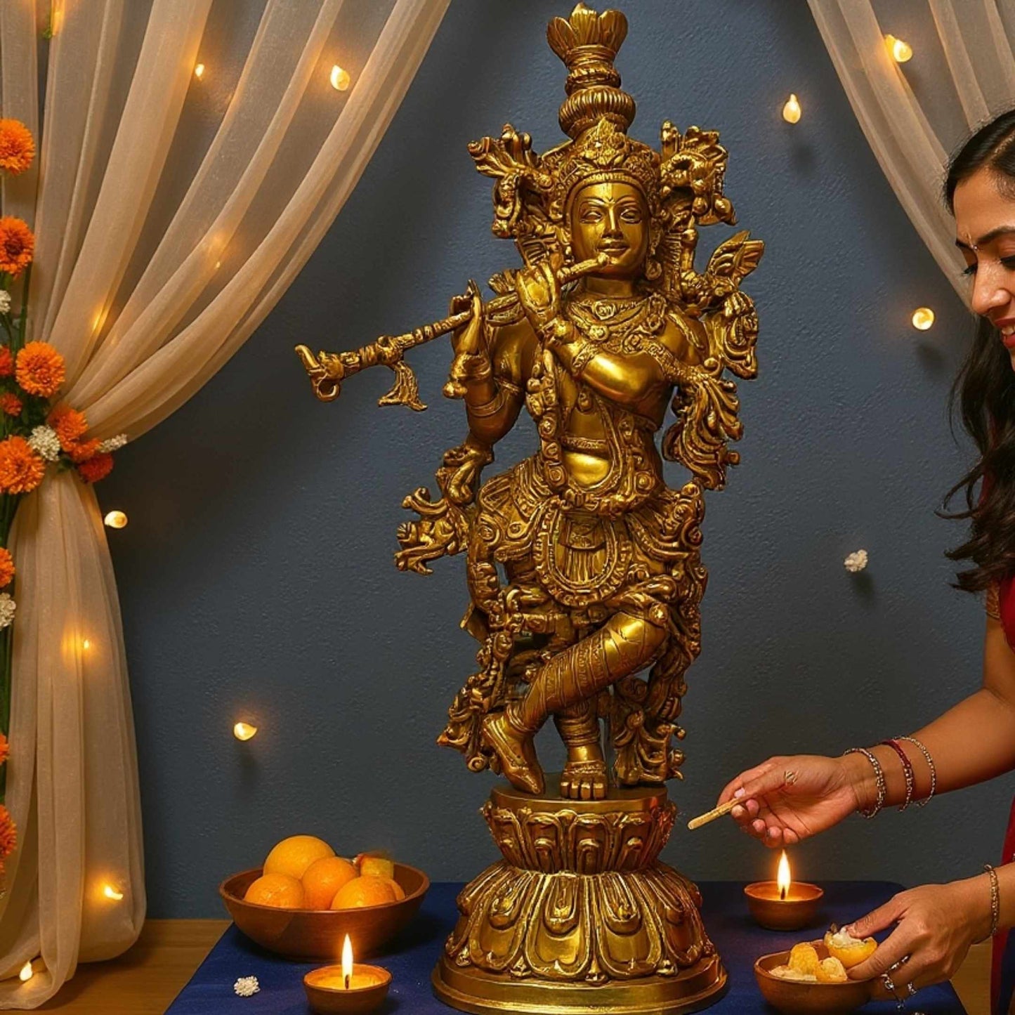 Brass Krishna statue displayed on a blue cloth in a decorated room with lit candles, fruit and flowers, as a woman lights a candle to the right
