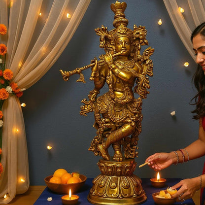 Brass Krishna statue displayed on a blue cloth in a decorated room with lit candles, fruit and flowers, as a woman lights a candle to the right