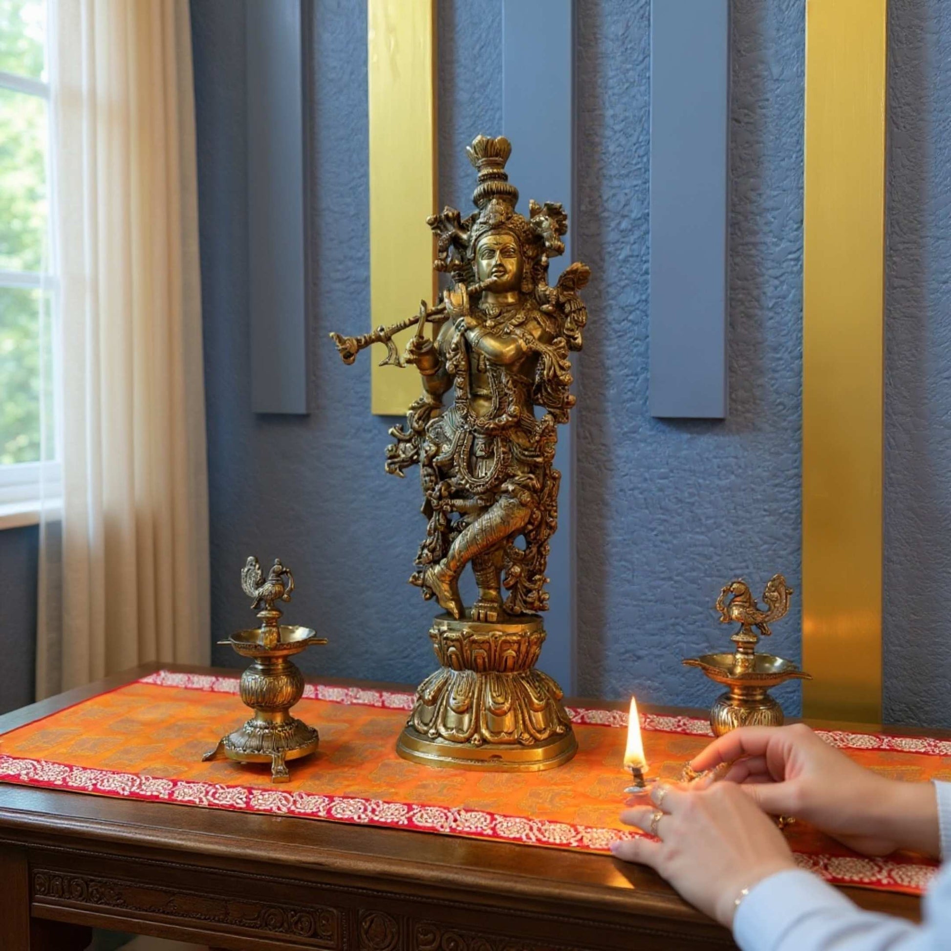 Brass Krishna statue on a decorated table with hands lighting a small oil lamp beside it, against a blue wall with gold vertical panels