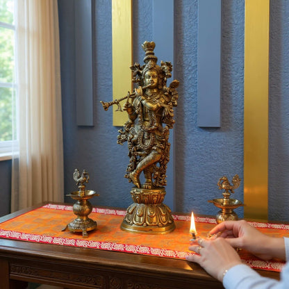 Brass Krishna statue on a decorated table with hands lighting a small oil lamp beside it, against a blue wall with gold vertical panels