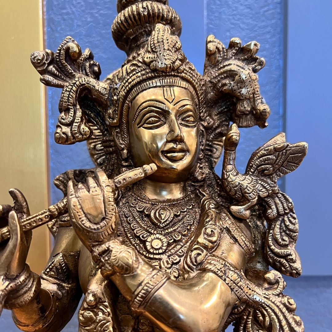 Brass Krishna statue playing the bansuri flute, showing the crown, jewellery and flanking scrollwork, against a blue textured wall