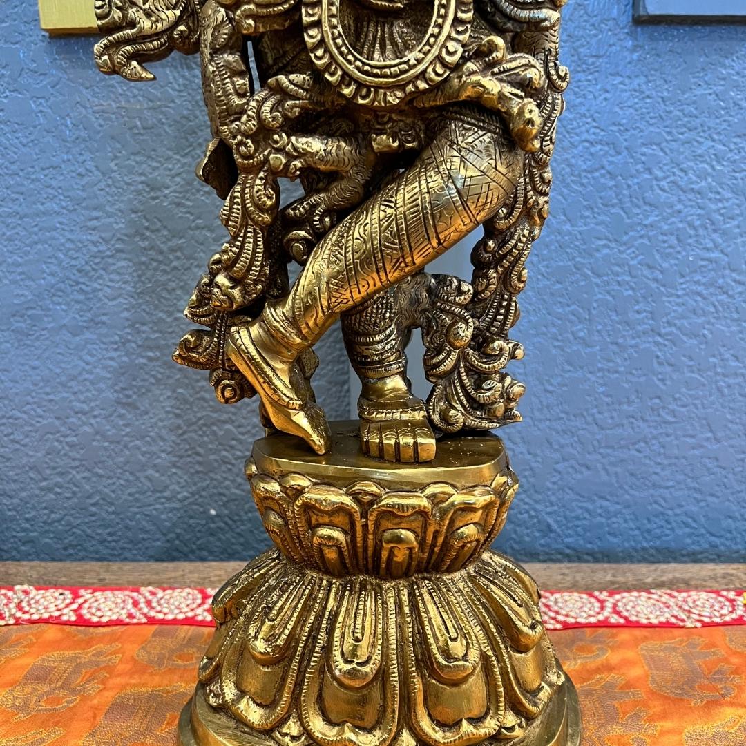 Close-up of the brass Krishna statue standing in tribhanga posture, showing the engraved dhoti and foliate scrollwork, against a blue wall and patterned surface