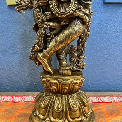 Close-up of the brass Krishna statue standing in tribhanga posture, showing the engraved dhoti and foliate scrollwork, against a blue wall and patterned surface