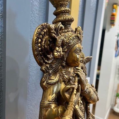 Close-up of the brass Krishna statue showing hand-engraved necklaces, armbands and the layered detailing of the crown, against a gray wall
