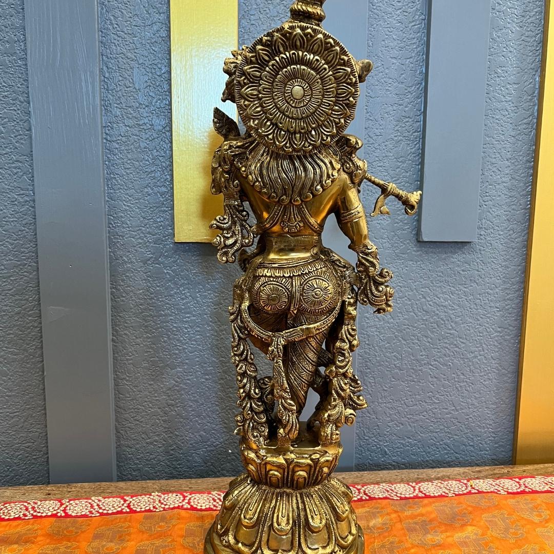 Back view of the brass Krishna statue showing the engraved lotus-mandala halo disc, layered hair and rosette-patterned dhoti, on its lotus pedestal against a blue-gray paneled wall