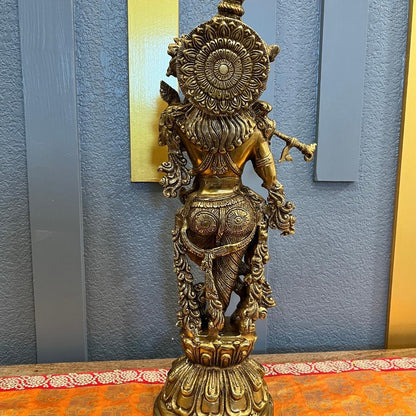 Back view of the brass Krishna statue showing the engraved lotus-mandala halo disc, layered hair and rosette-patterned dhoti, on its lotus pedestal against a blue-gray paneled wall