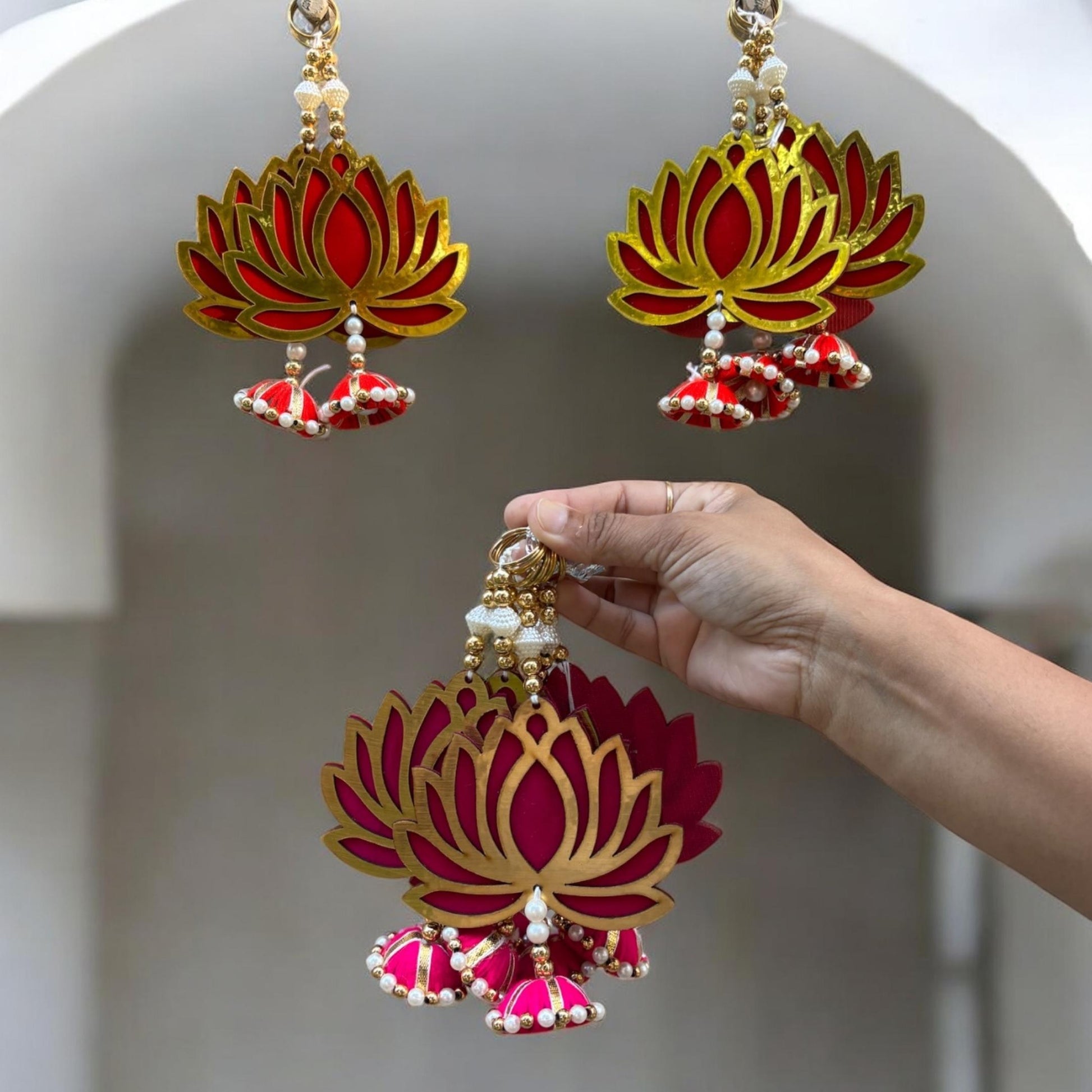 Lotus-shaped danglers with red, gold, and white colors held by a hand against a neutral background.