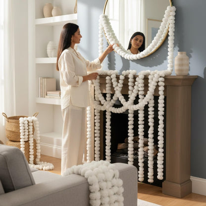 Woman arranging decorative white marigold garlands on a fireplace mantel in a living room.