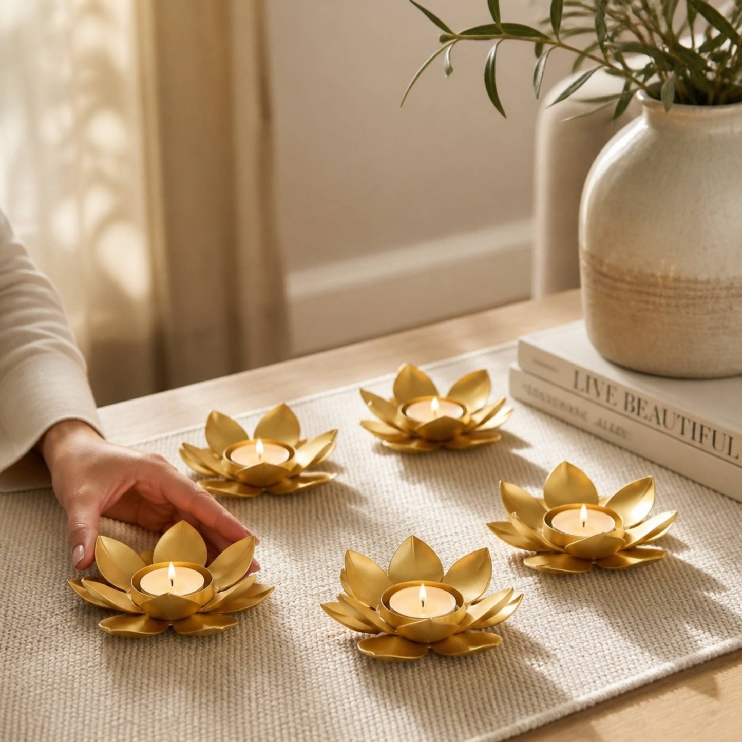 Five gold lotus t-light holders with lit tea lights on a woven table runner near a vase and books