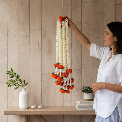 Woman arranging a decorative garland on a wooden wall with a table and plants in the foreground.