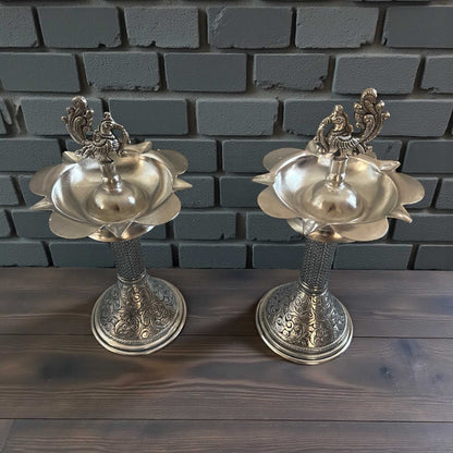 Two ornate silver samai oil lamp stands on wooden surface with brick background
