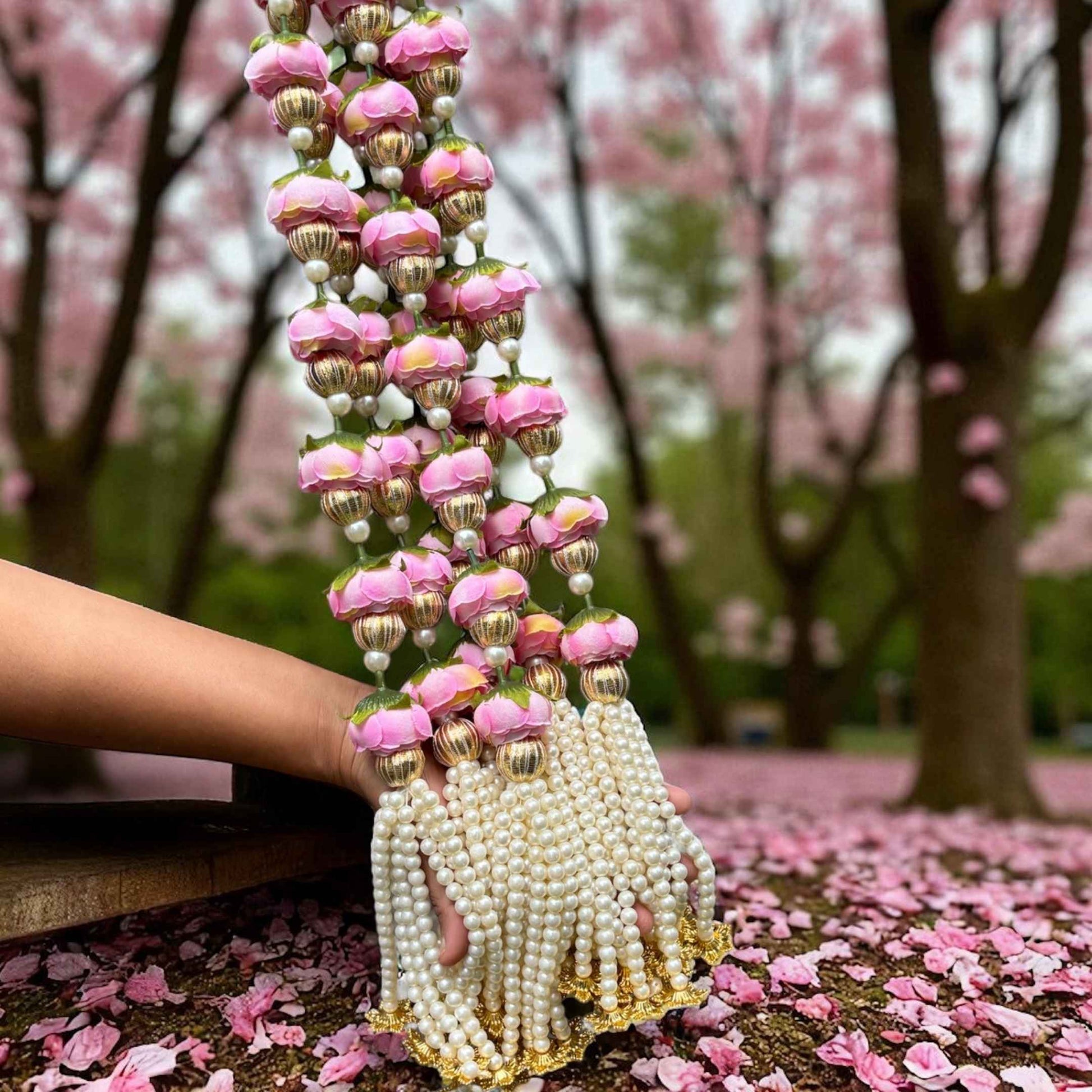 Decorative garland with pink flowers and pearls held by a hand on a ground covered with pink petals, with cherry blossom trees in the background.