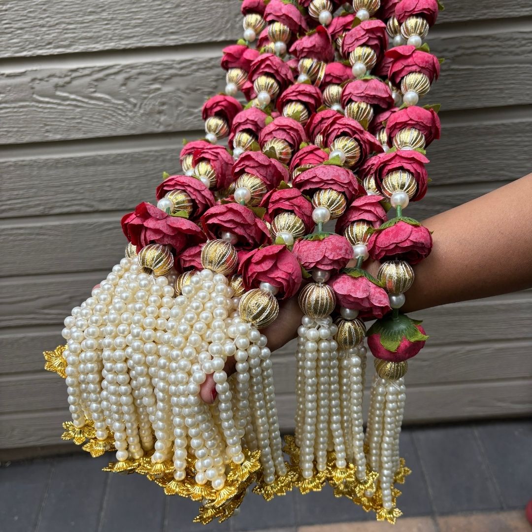 Decorative garland with red flowers, gold balls, and white pearls held by a person against a wooden wall.