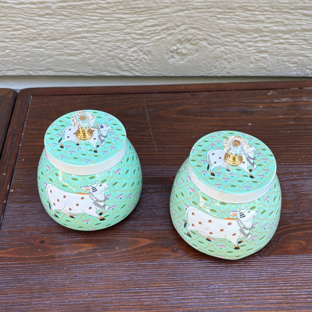 Two decorative jars with pichwai cow illustrations on a wooden surface.