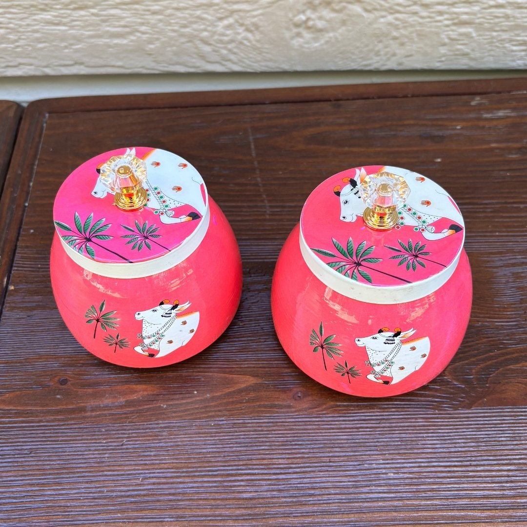 Two pink metal jars with decorative lids on a wooden surface.