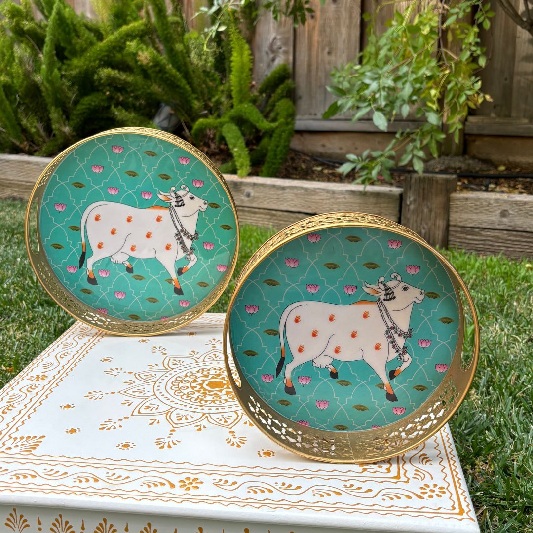 Two circular mint metal trays painted with a white Pichwai-style cow motif, displayed upright against greenery to showcase design and color contrast.