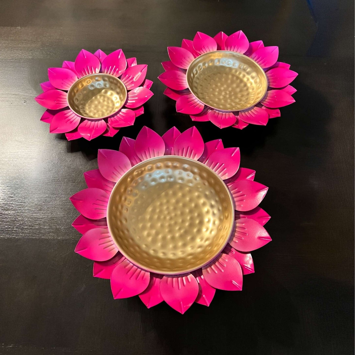 Three pink flower-shaped candle holders urli bowls with gold centers on a dark surface
