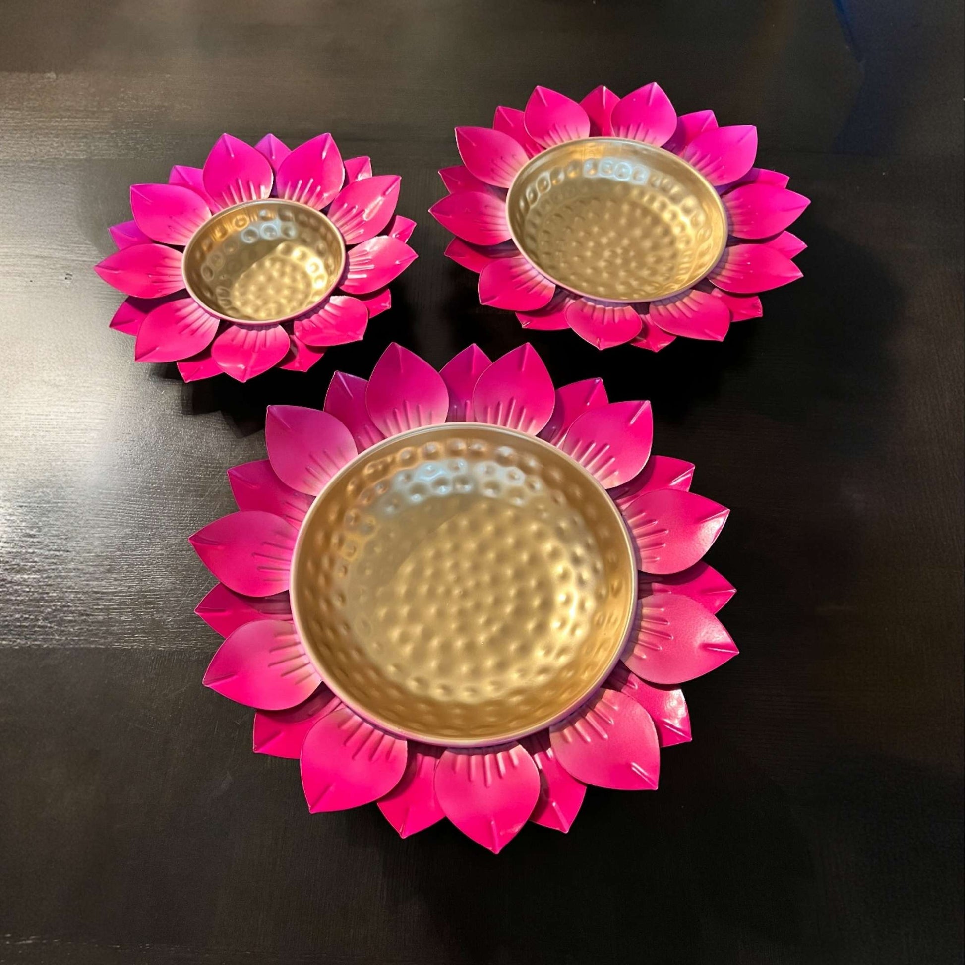 Three pink flower-shaped candle holders urli bowls with gold centers on a dark surface