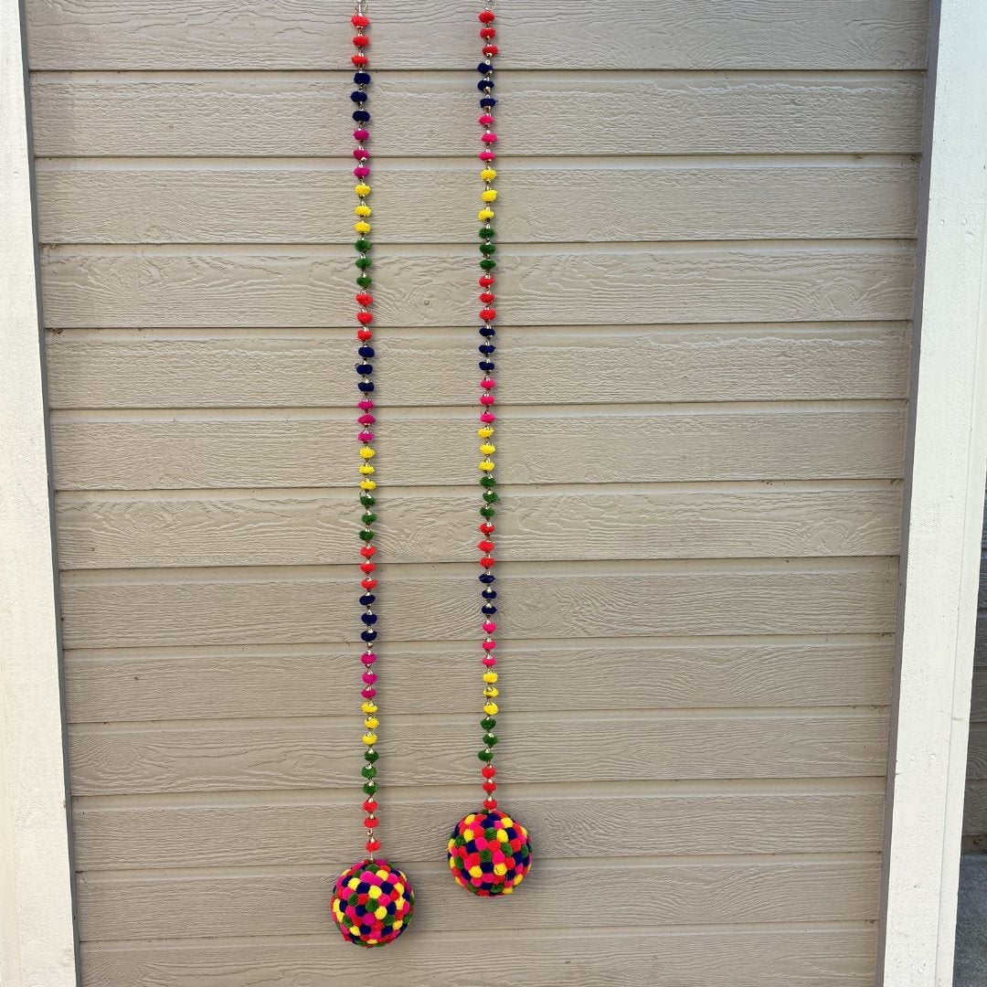 Two long beaded hanging garlands with colorful round pom-poms, displayed against a beige horizontal siding wall.