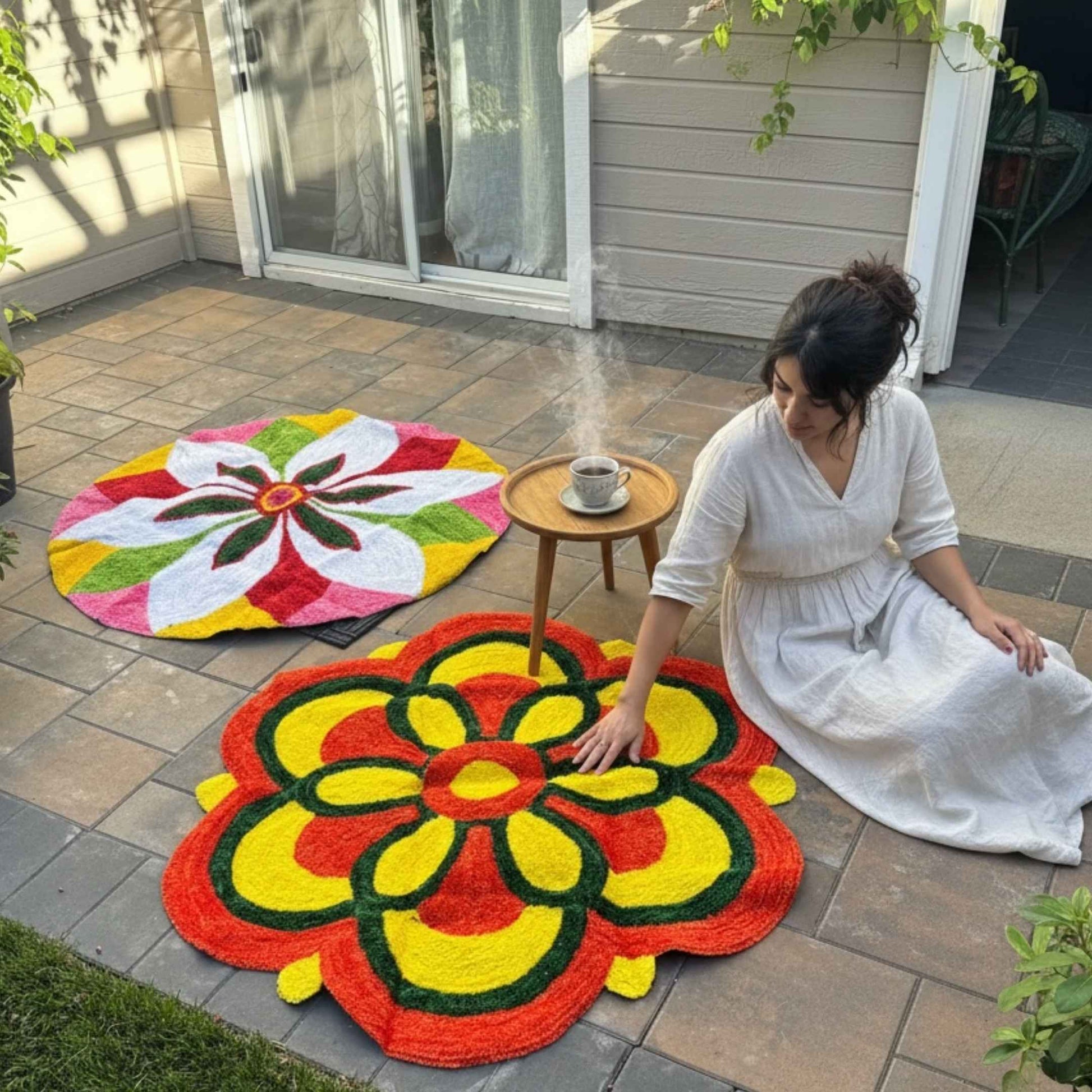 Two rangoli mats on a patio, woman in white sitting beside a yellow-and-red mat with steaming tea on a side table