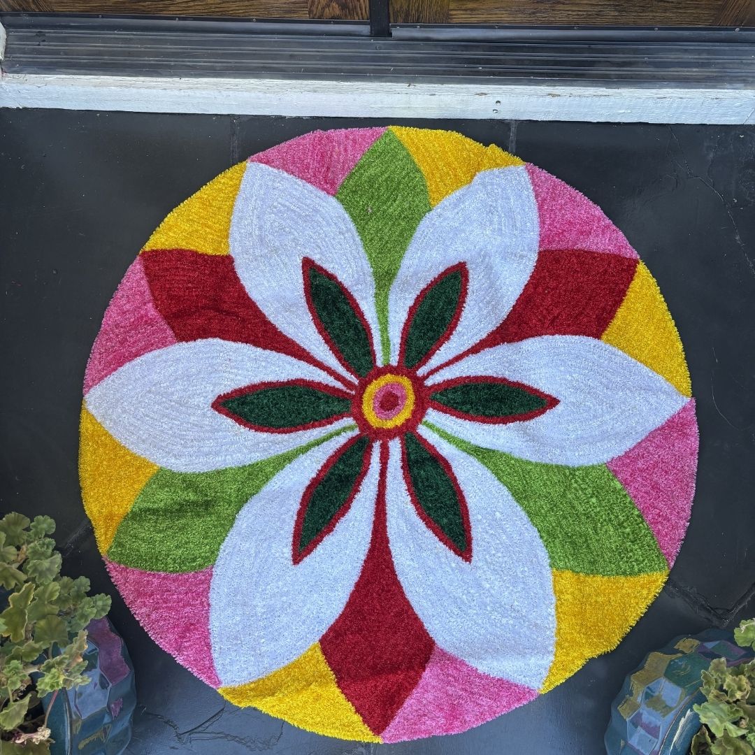 Round rangoli mat with a white flower design in green, red, pink, and yellow segments, indoors