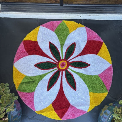 Round rangoli mat with a white flower design in green, red, pink, and yellow segments, indoors