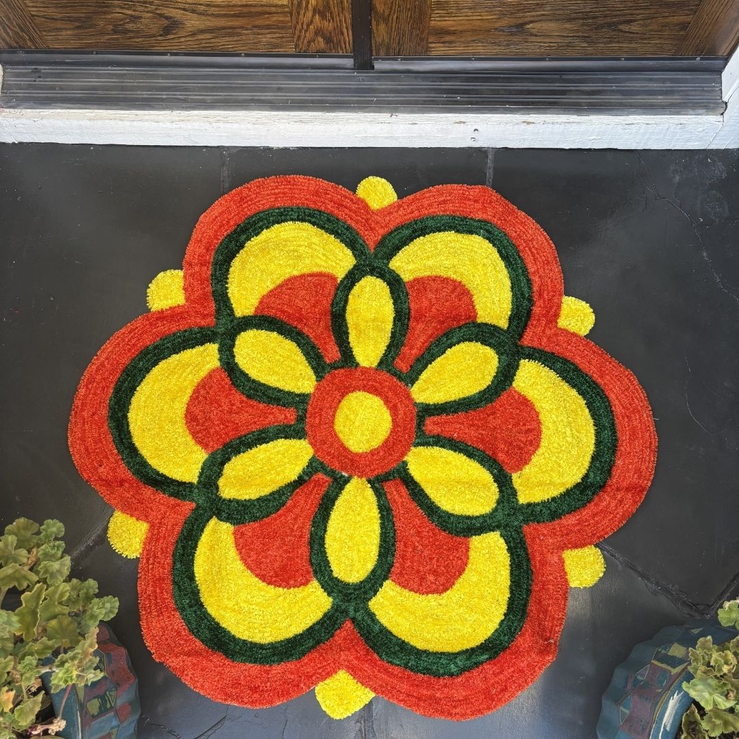 Round rangoli mat with a yellow flower center and a green, orange, and yellow scalloped petal border, indoors