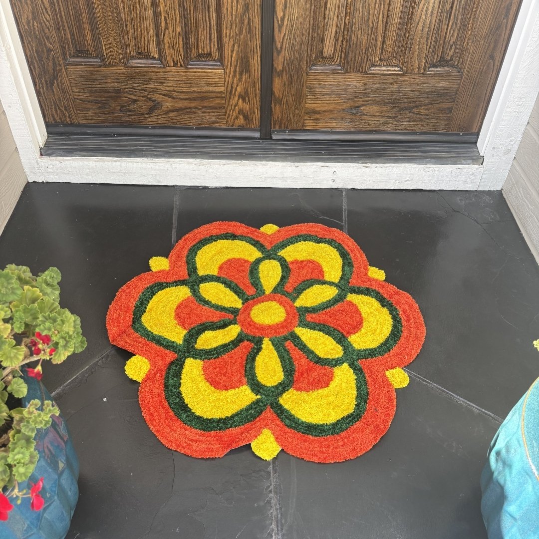 Small round rangoli mat with a yellow flower center and orange scalloped border on a dark tile entryway