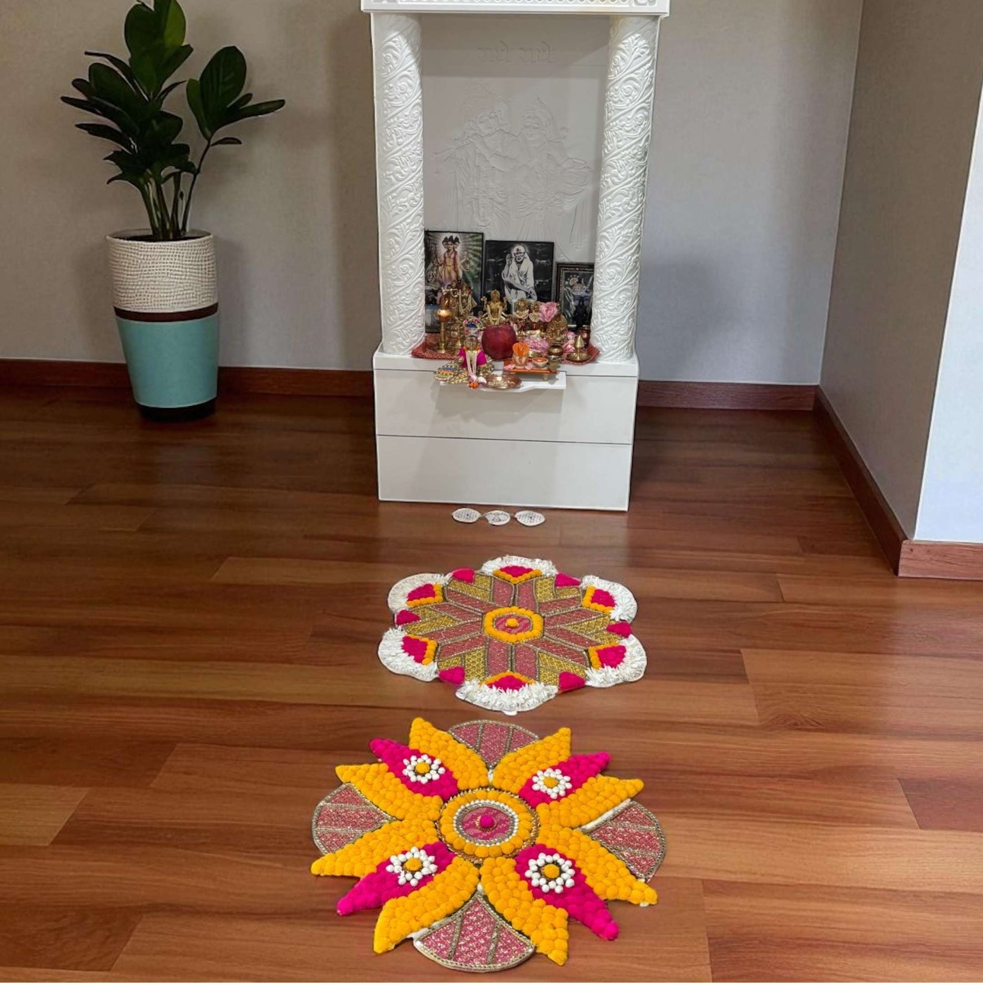 Decorative rangoli mat arrangements on a wooden floor with a white pedestal and plant in the background.