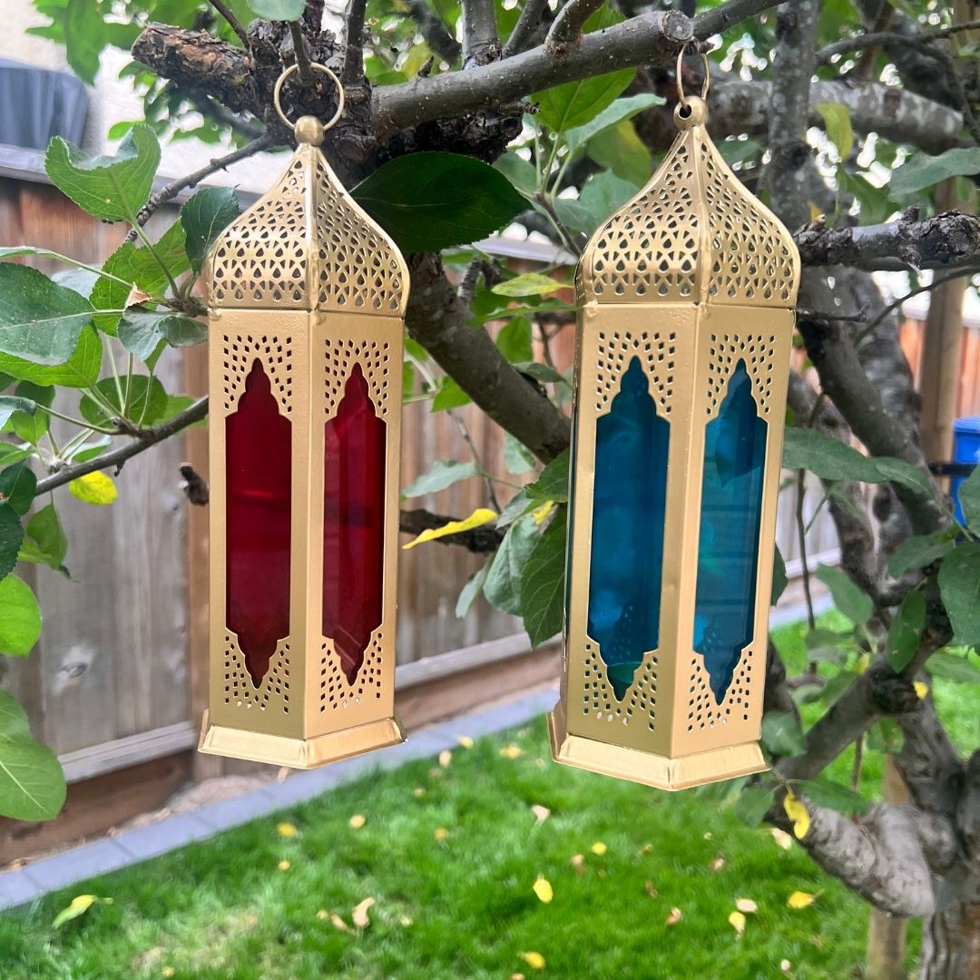 Handcrafted Moroccan-style tealight lanterns with red and blue stained glass panels, suspended outdoors, casting vibrant hues—ideal for garden or patio décor.
