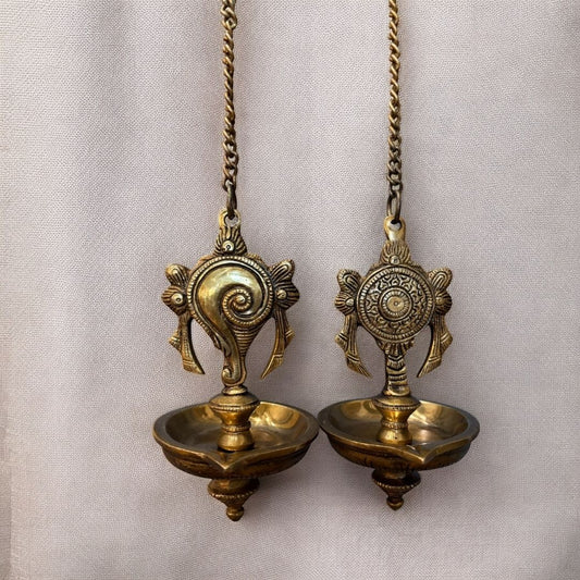 Two ornate brass hanging diya lamps suspended from chains against a light-colored background; each lamp features a decorative shankh-and-chakra motif above a lotus-shaped bowl.