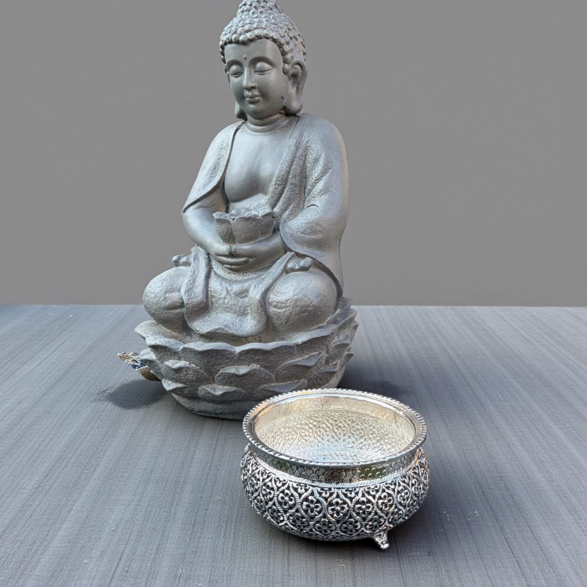 Antique silver engraved urli bowl placed beside Buddha statue as home decor vignette, german silver finish textured scene