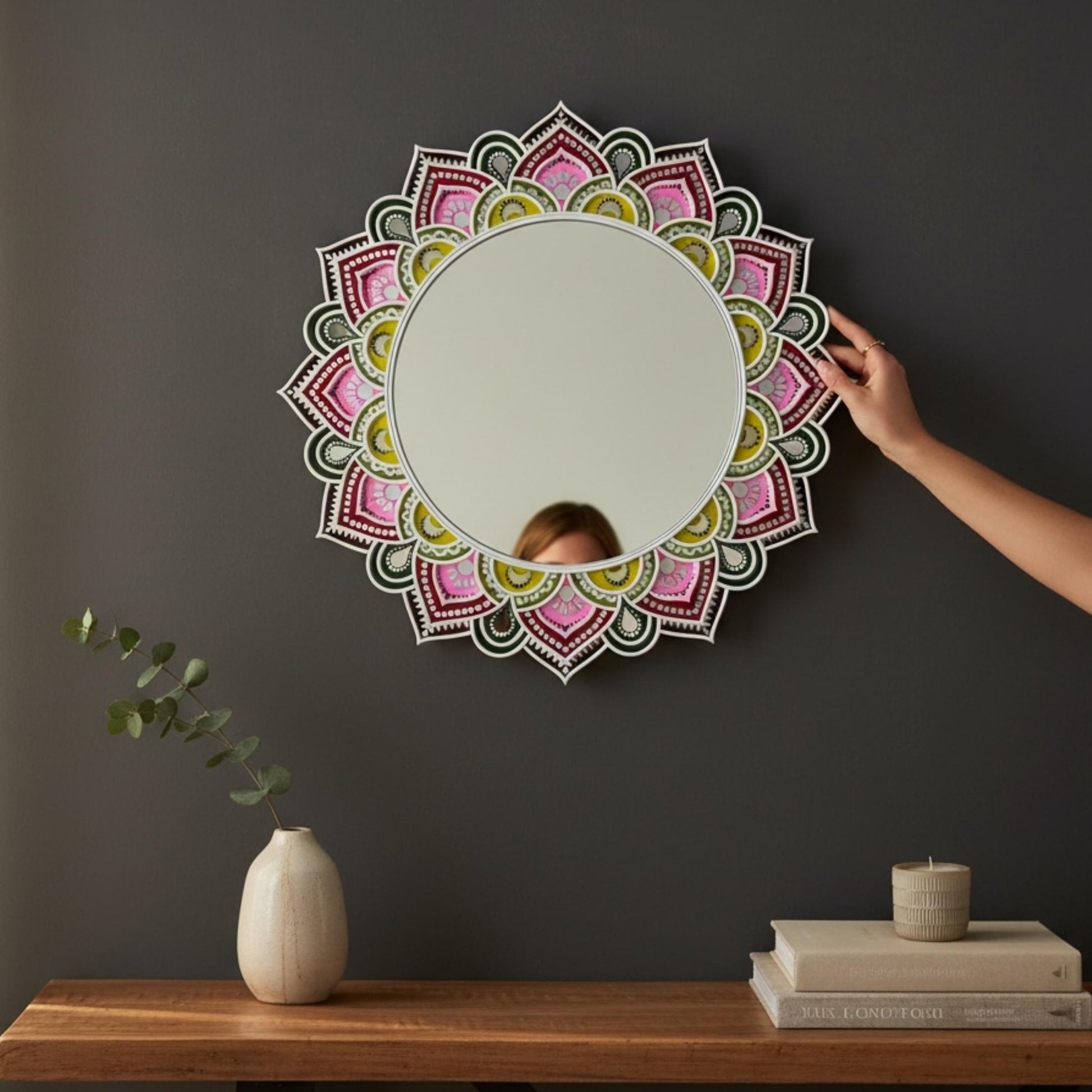 Decorative lippan mirror with mandala design on a dark wall, with a hand adjusting it.