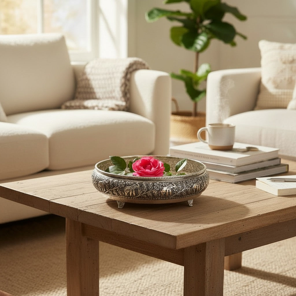 Antique silver elephant urli bowl holding a pink rose and leaves on a wooden coffee table in a bright living room