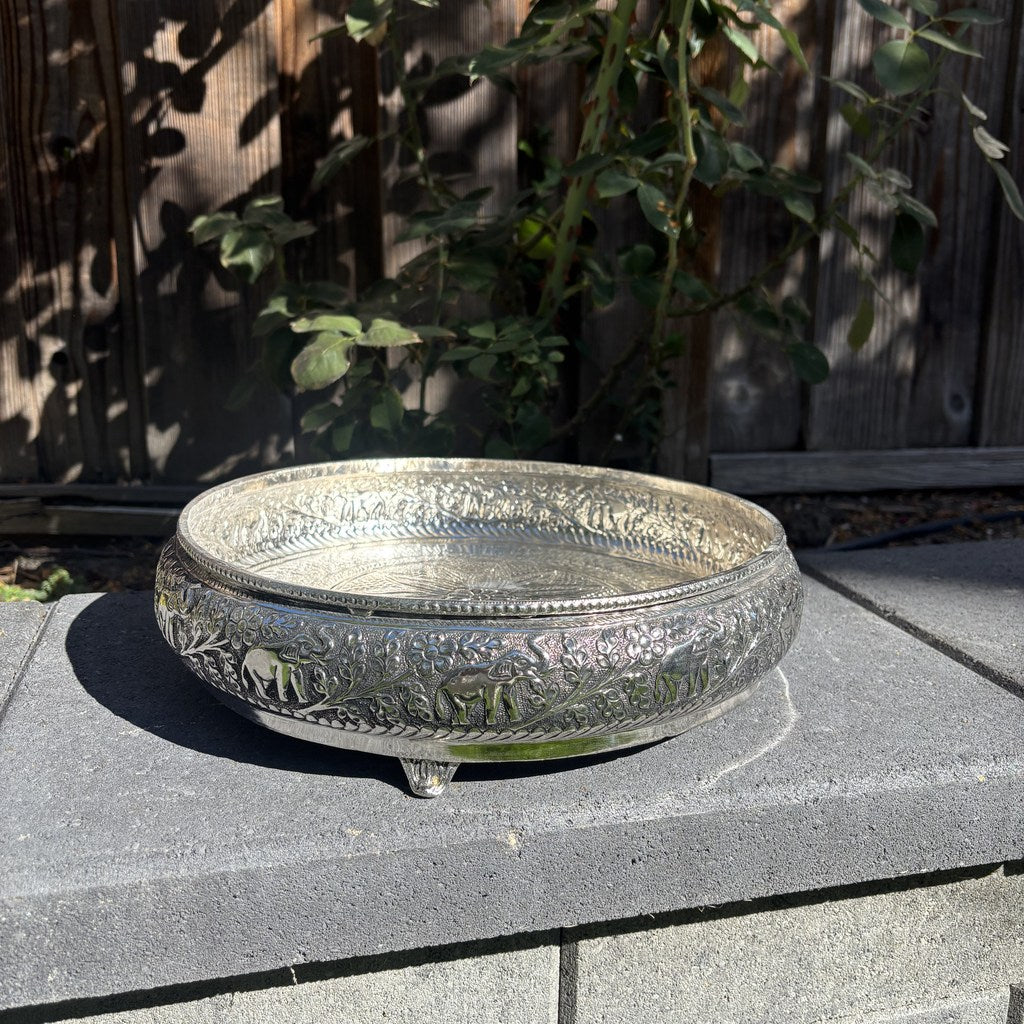 Side view of the antique silver urli bowl on its small feet, showing raised elephants and vines around the wall