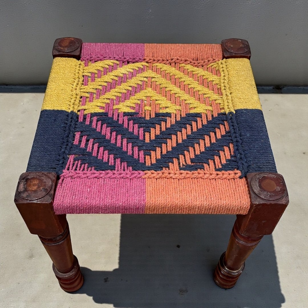 Small stool with colorful woven top and wooden legs on a concrete floor.