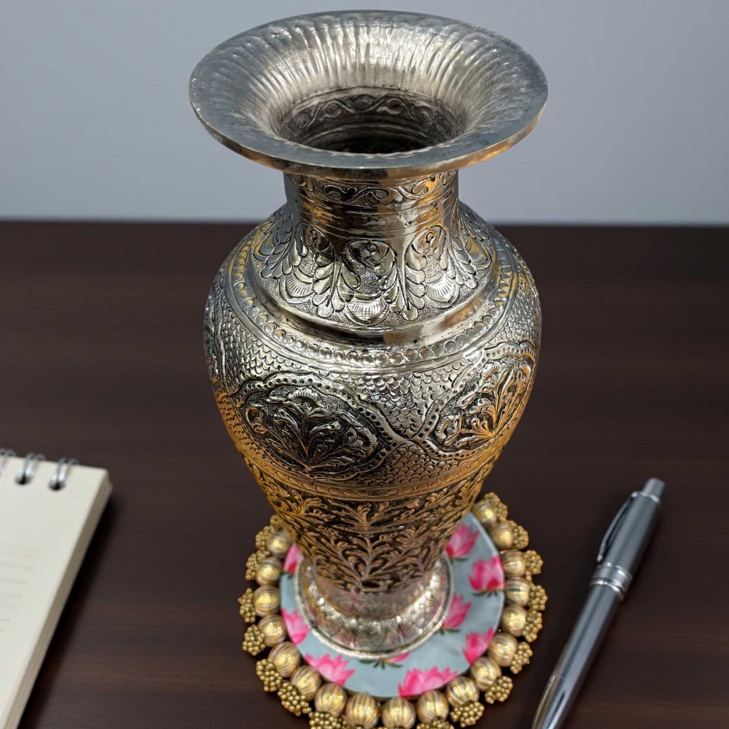 Decorative silver vase with intricate designs on a wooden surface with a pen and notebook.
